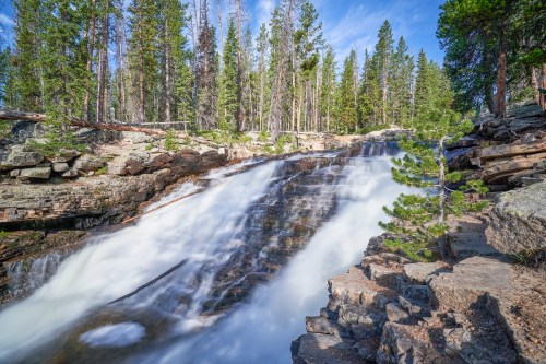 A serene waterfall cascades over rocky terrain, surrounded by lush green trees under a clear blue sky.