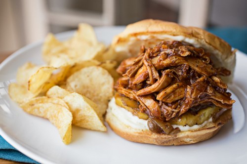 A pulled chicken sandwich topped with barbecue sauce, served with a side of crispy potato chips on a white plate.