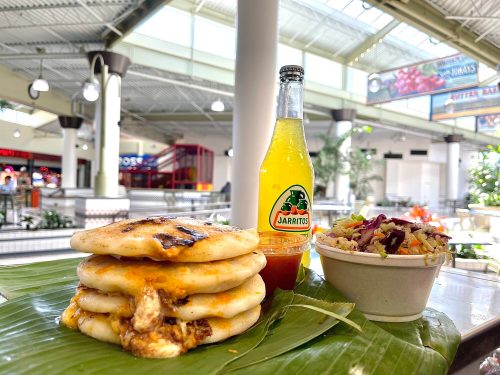 A stack of grilled tortillas with filling, a bottle of Jarritos soda, and a bowl of colorful coleslaw on a banana leaf.