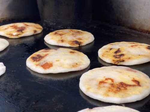 A close-up of golden-brown pupusas cooking on a griddle, with a slightly charred surface.