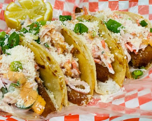 Four delicious tacos topped with creamy sauce, cabbage, green onions, and crumbled cheese, served with a lemon wedge.