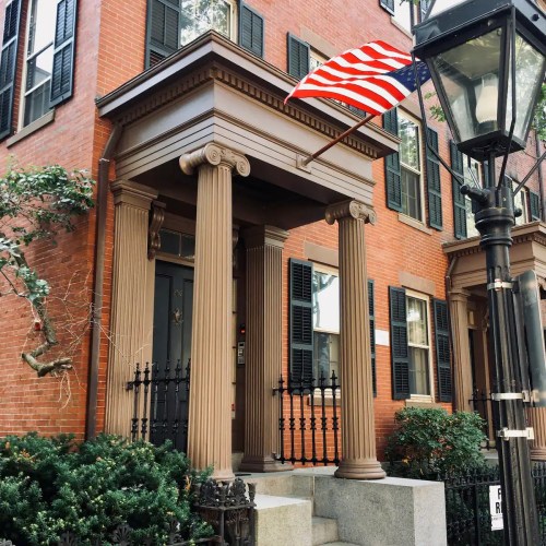 A historic brick building with a columned entrance, an American flag, and a lamppost nearby.