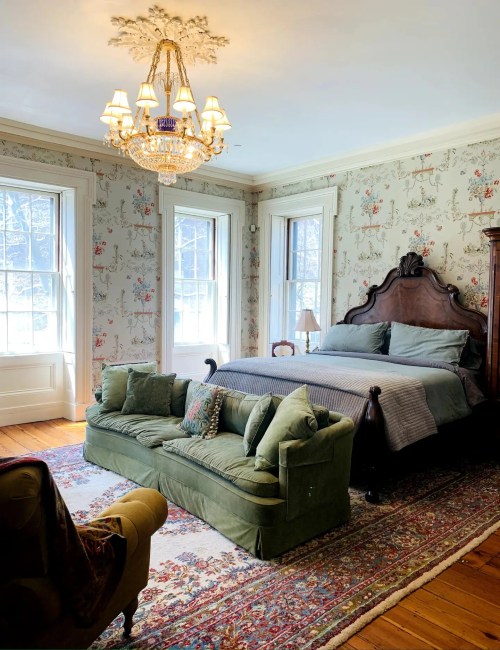 A cozy bedroom featuring a large bed, green sofa, chandelier, and floral wallpaper with large windows.