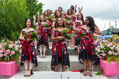 A group of smiling women in floral dresses holding bouquets, standing on steps decorated with flowers.