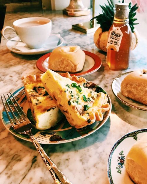 A plate with a slice of quiche, pastries, a cup of tea, and a bottle of honey on a marble table.