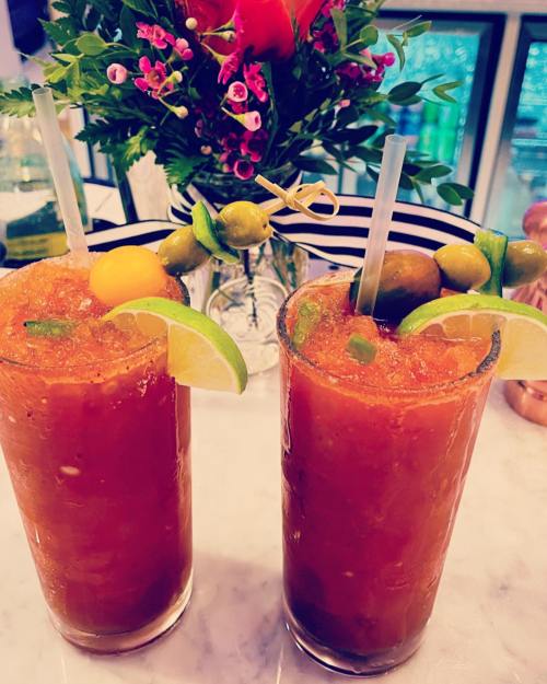 Two tall glasses of vibrant Bloody Mary cocktails, garnished with olives, lime, and a cherry, on a marble countertop.