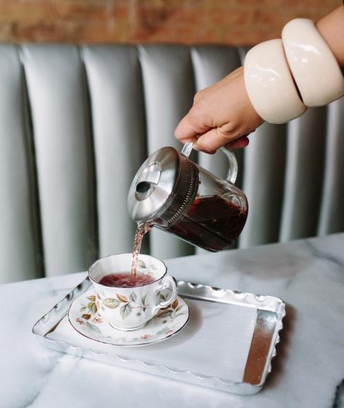 A hand pours tea from a glass teapot into a delicate floral teacup on a silver tray.