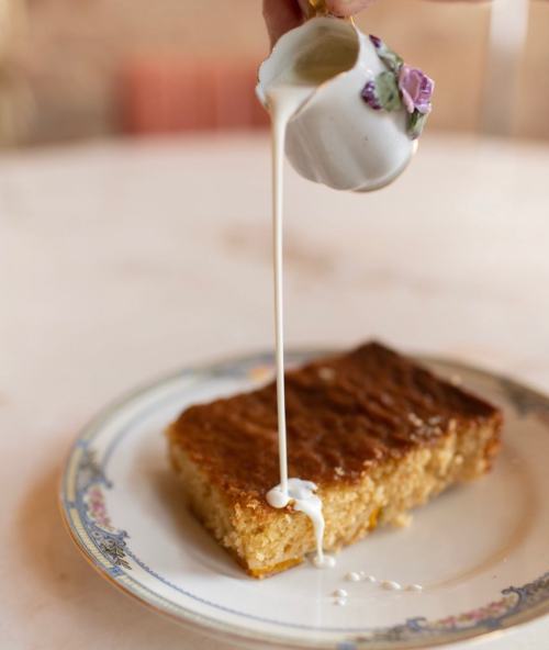 A hand pours cream from a delicate cup onto a slice of golden cake on a decorative plate.