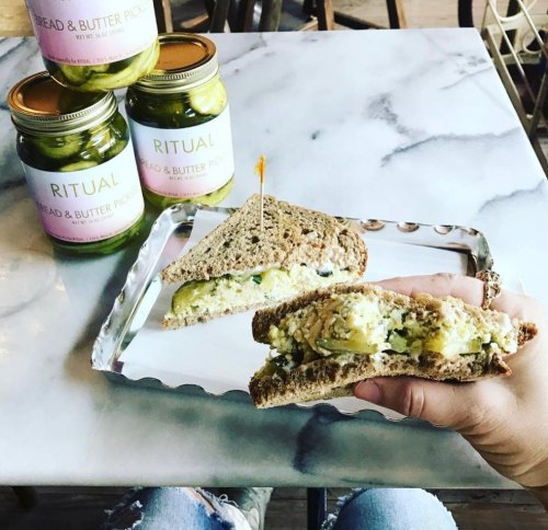 A hand holds a sandwich on a plate, with jars of pickles in the background on a marble table.