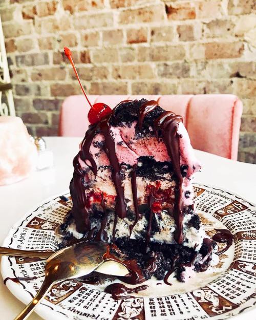 A slice of layered ice cream cake topped with chocolate drizzle and a cherry, served on a decorative plate.
