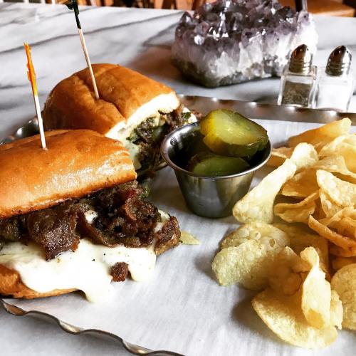 Two sandwiches with toppings, a side of pickles, and a plate of potato chips on a table.