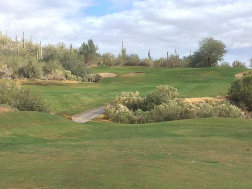 A lush green golf course with rolling hills, sand traps, and desert vegetation under a cloudy sky.