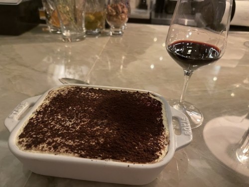 A serving of tiramisu in a white dish next to a glass of red wine on a marble countertop.