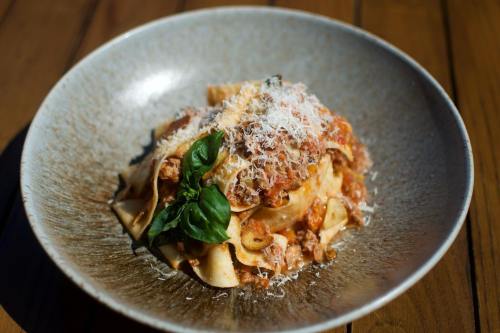 A bowl of pappardelle pasta topped with meat sauce, grated cheese, and a fresh basil leaf on a wooden table.