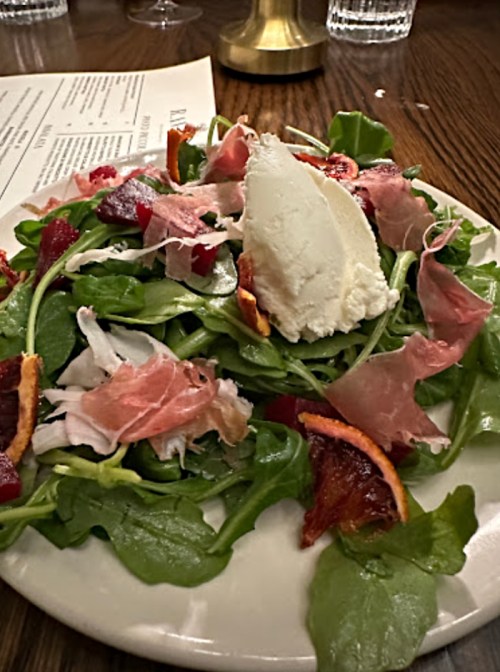 A fresh salad with greens, prosciutto, shredded chicken, dried fruit, and a dollop of creamy cheese on top.