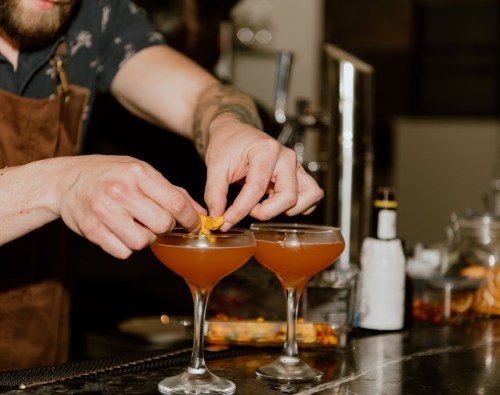 A bartender garnishes two cocktails with a twist of citrus at a stylish bar.