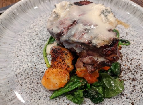 A plated dish featuring a piece of meat topped with creamy sauce, served on a bed of spinach and gnocchi with vegetables.