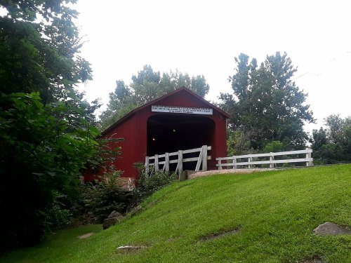 A red covered bridge surrounded by greenery and trees, with a white fence in front.