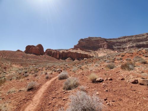 A winding dirt path leads through a rocky, arid landscape with red hills and sparse vegetation under a clear blue sky.
