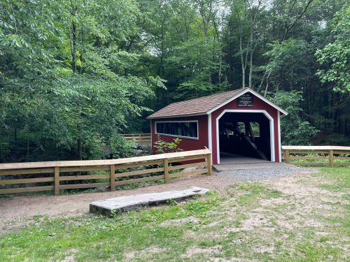 A red covered bridge surrounded by lush green trees and a wooden fence in a serene outdoor setting.