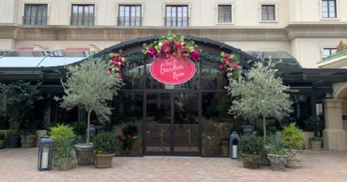 Entrance to "The Garden Room," featuring floral decorations and greenery, with a glass canopy and potted plants.