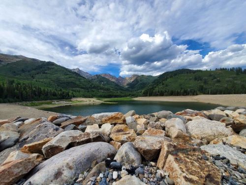 A serene lake surrounded by green mountains and a cloudy sky, with a rocky shoreline in the foreground.