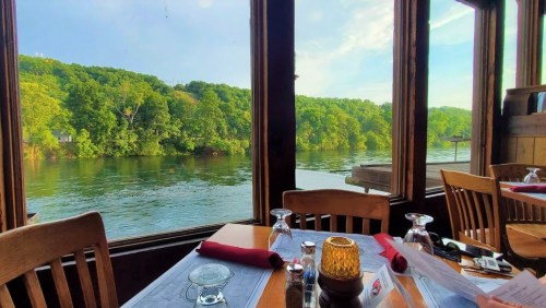 A scenic view from a restaurant window overlooking a river and lush green hills. Tables are set for dining.