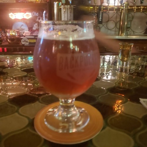 A glass of amber beer sits on a bar counter, with a blurred background of bottles and bar lights.