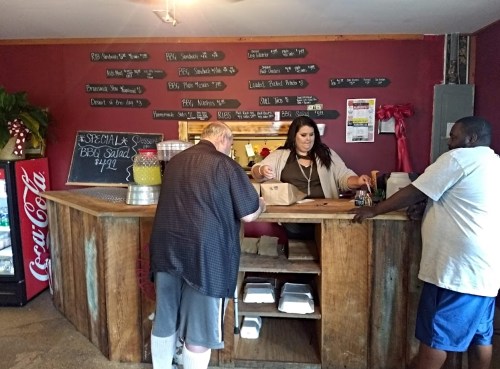 A woman serves customers at a rustic counter in a BBQ restaurant, with a menu displayed on the wall behind her.