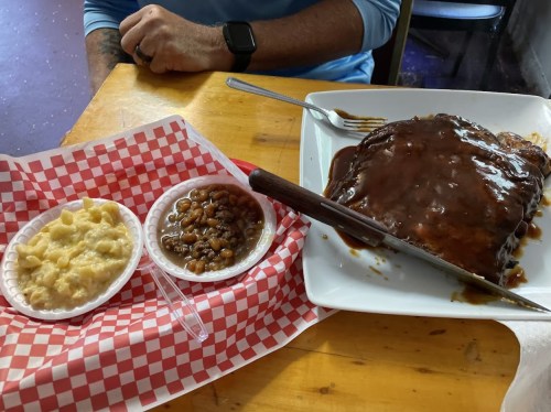 A plate with a large serving of meat covered in sauce, accompanied by sides of macaroni and cheese and baked beans.