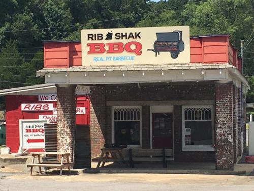 A rustic BBQ restaurant with a sign reading "Rib Shak BBQ" and picnic tables outside, surrounded by greenery.