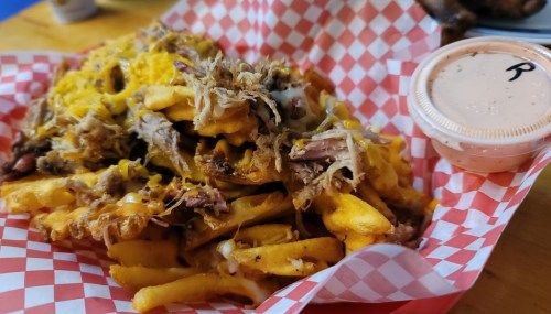 A basket of loaded fries topped with pulled pork and cheese, accompanied by a small container of sauce.