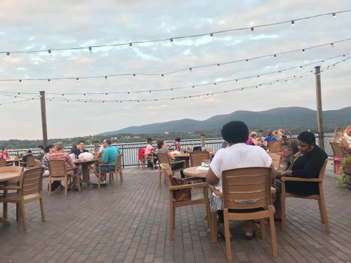 Outdoor dining area with people at tables, string lights overhead, and mountains in the background under a cloudy sky.