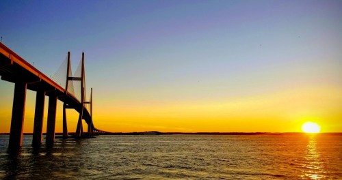 A bridge spans over water at sunset, with vibrant orange and yellow hues illuminating the sky.