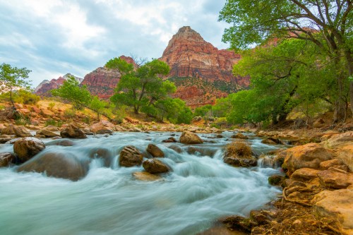 A serene river flows over rocks, surrounded by lush greenery and towering red rock formations under a cloudy sky.