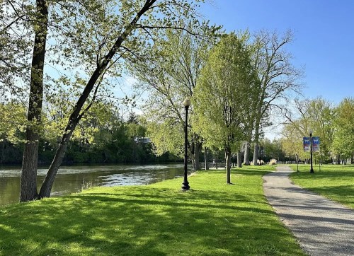 A serene riverside park with green grass, trees, and a walking path under a clear blue sky.