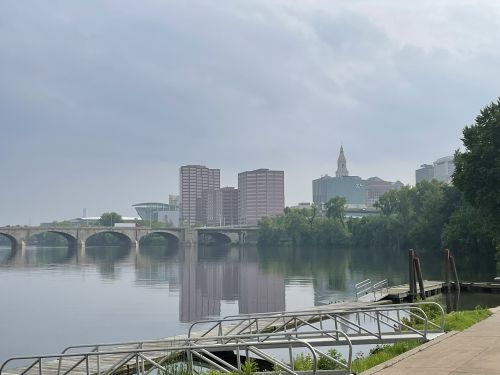 A calm river reflects a city skyline with buildings and a bridge under a cloudy sky.