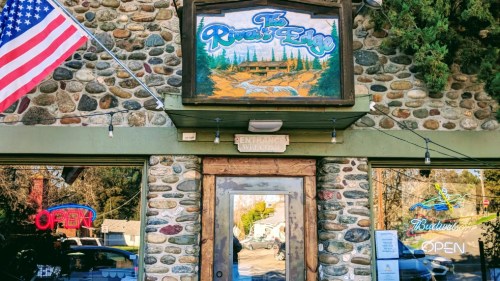 A rustic stone building with a colorful sign reading "The River's Edge" and an open door, featuring an American flag.