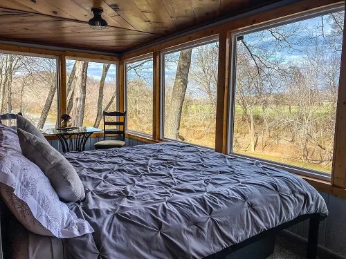 Cozy bedroom with a large bed and windows overlooking a serene outdoor view of trees and fields.