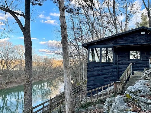A dark wooden cabin by a river, surrounded by trees and a clear blue sky with scattered clouds.