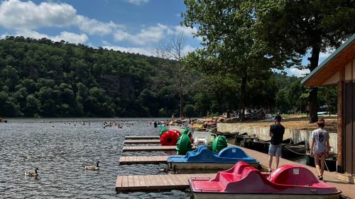 A serene lake scene with colorful paddle boats lined up on a dock, surrounded by trees and people enjoying the water.