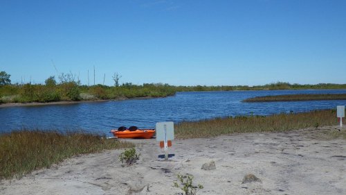 A calm waterway with an orange kayak on the shore, surrounded by green vegetation and a clear blue sky.