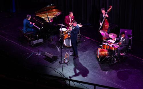 A jazz band performs on stage, featuring a pianist, trumpeter, drummer, and bassist, with dramatic lighting and shadows.