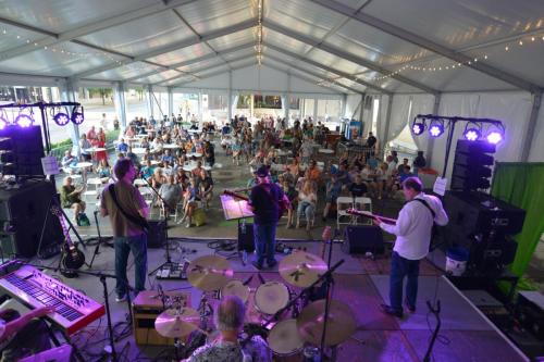 A band performs on stage inside a tent, with an audience seated and enjoying the music.