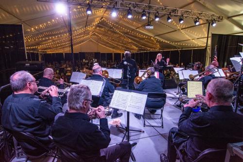 A conductor leads an orchestra under a tent with string lights, while musicians play instruments on stage.