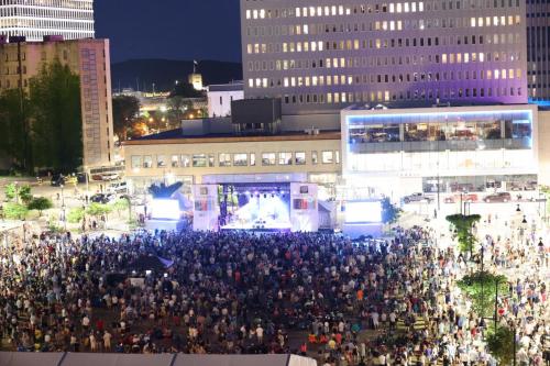 A large crowd gathers at night for a concert in an urban setting, with bright stage lights and buildings in the background.