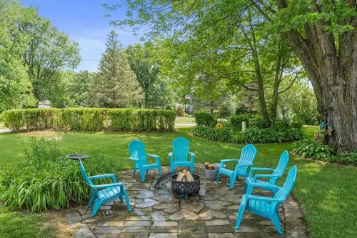 A cozy outdoor seating area with turquoise chairs around a fire pit, surrounded by lush greenery and trees.