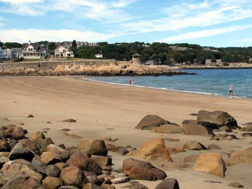 A sandy beach with scattered rocks, people walking along the shore, and houses on a hillside in the background.