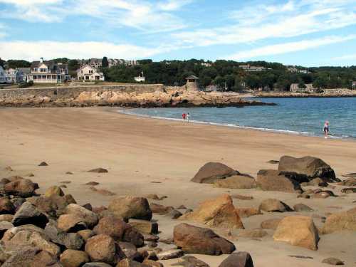 A sandy beach with scattered rocks, people walking along the shore, and houses on a hillside in the background.