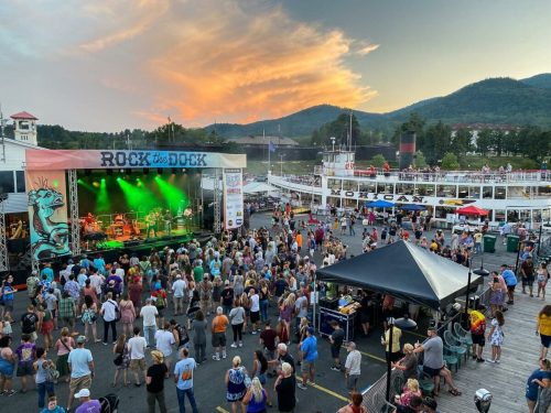 A lively outdoor concert at sunset, with a large crowd enjoying music near a dock and scenic mountains in the background.